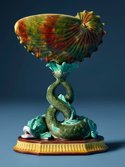 Majolica centerpiece featuring a nautilus shell bowl supported by intertwining fish, set against a soft blue background.