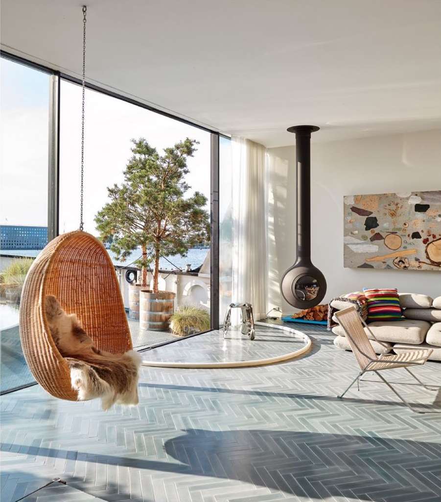Indoor living room featuring a hanging wicker chair with fur throw, modern fireplace, and large windows overlooking greenery.