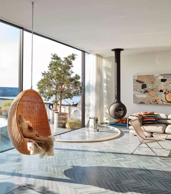 Indoor living room featuring a hanging wicker chair with fur throw, modern fireplace, and large windows overlooking greenery.