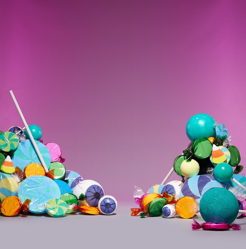 Colorful candy piles on a gray surface against a graduated magenta background, featuring lollipops, wrapped candies, and eyeball candies.