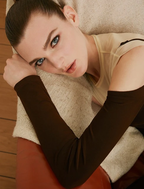 Close-up of a woman with blue eyes lying down indoors, wearing a beige top and long brown sleeves, resting on a cream colored cushion.