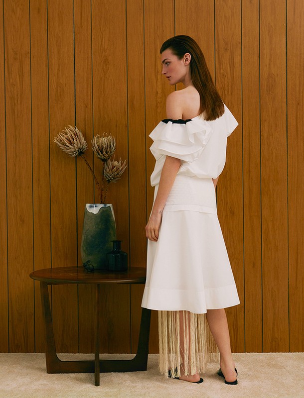 Model wearing a white one-shoulder ruffled dress standing beside a wooden table with a ceramic vase in Los Angeles.