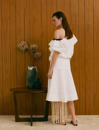 Model wearing a white one-shoulder ruffled dress standing beside a wooden table with a ceramic vase in Los Angeles.