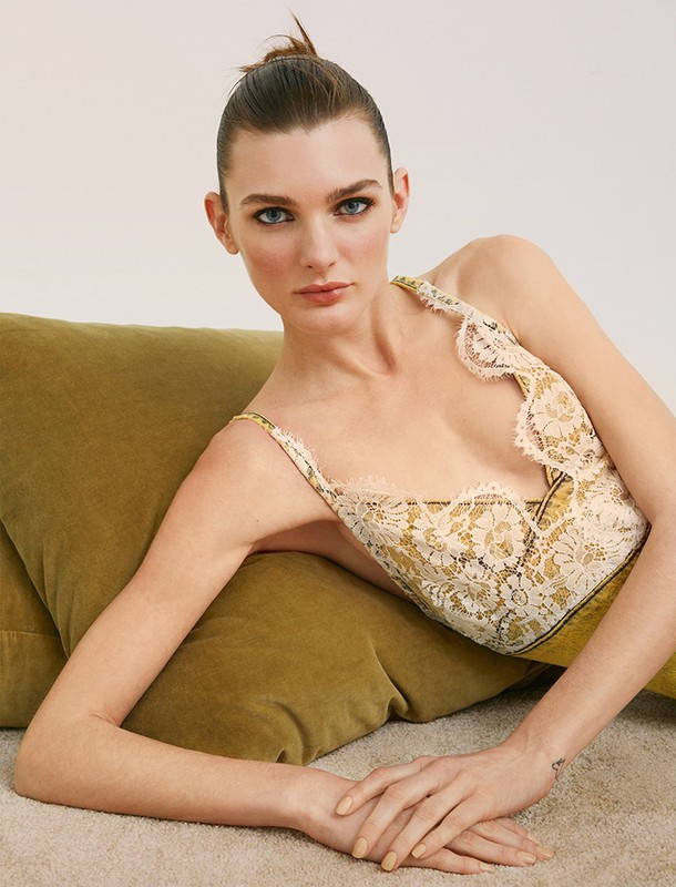 A woman in a lace camisole reclines on an olive pillow against a light background, looking directly at the camera in a studio shot.
