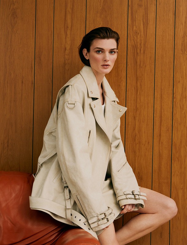 A stylish woman with short hair, wearing a tan trench coat, sits on a red-orange leather chair against a wooden wall in a studio setting.