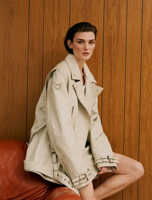 A stylish woman with short hair, wearing a tan trench coat, sits on a red-orange leather chair against a wooden wall in a studio setting.