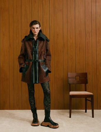 Model wearing a brown patterned coat with black trim and chunky boots standing against a wood-paneled wall with a chair.