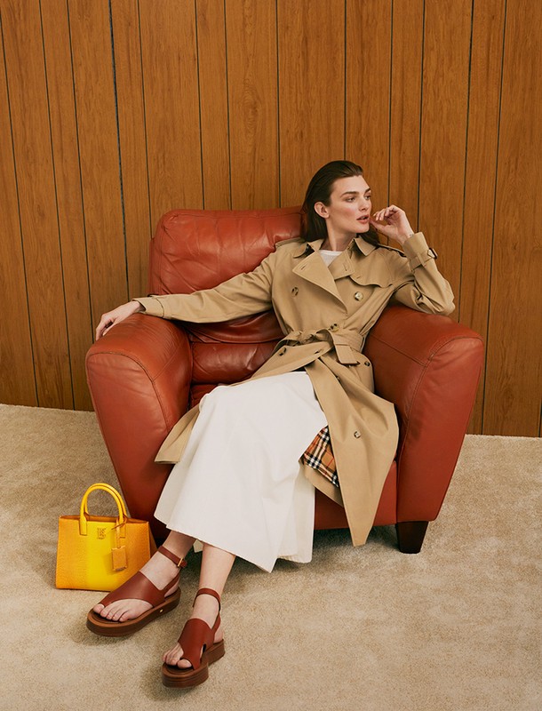 A woman in a trench coat sits in a red leather armchair indoors next to a yellow leather purse and brown sandals, fashion-forward, styled.