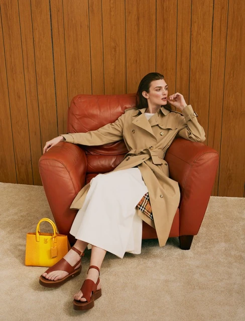 A woman in a trench coat sits in a red leather armchair indoors next to a yellow leather purse and brown sandals, fashion-forward, styled.