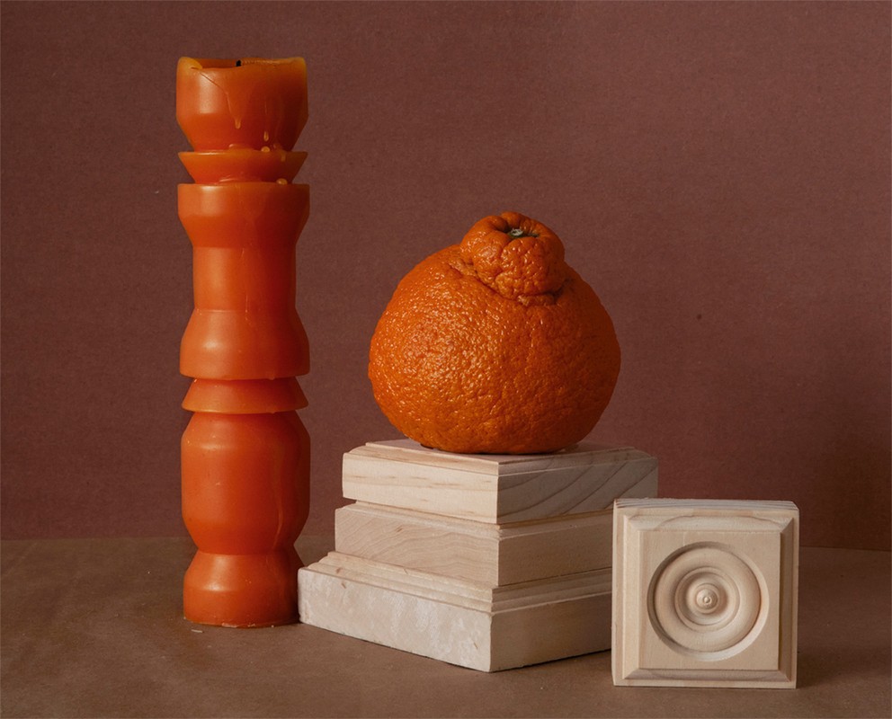Still life photograph features an orange citrus fruit next to an orange candle, set atop light-colored wood blocks against a brown backdrop.