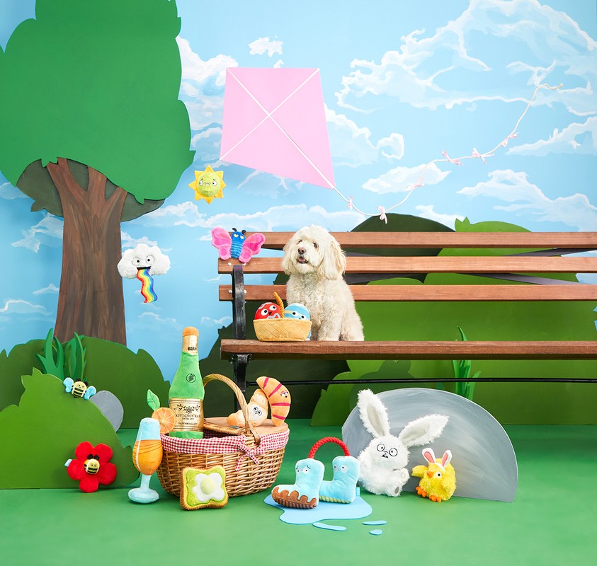 A white Goldendoodle dog sits on a wooden park bench with colorful toys, a picnic basket, and a kite against a sky-painted backdrop.