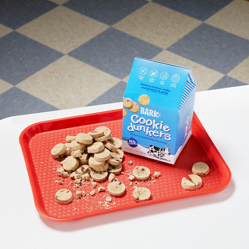 Pile of Bark Cookie Dunkers dog treats spilling from a light blue carton onto a red plastic tray, set on a white table.