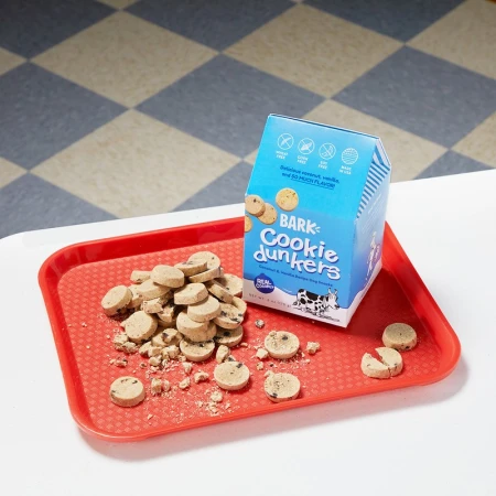 Pile of Bark Cookie Dunkers dog treats spilling from a light blue carton onto a red plastic tray, set on a white table.