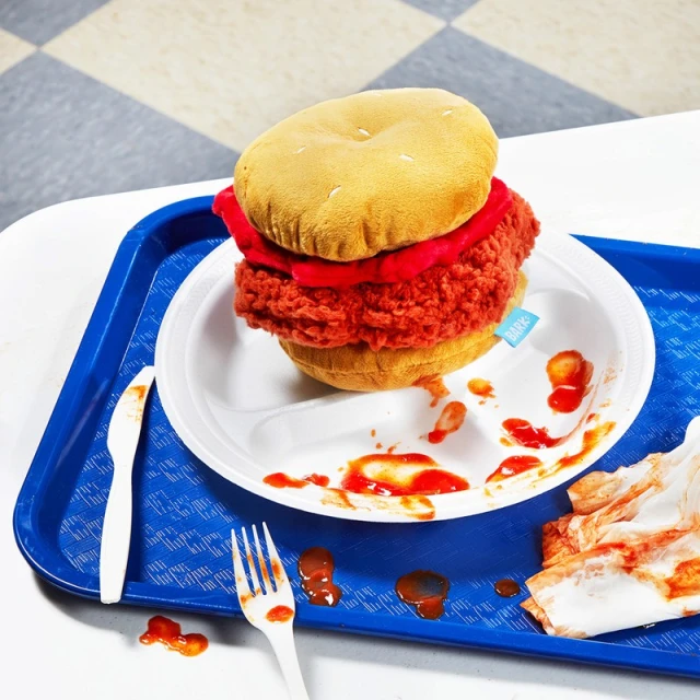 Plush toy chicken sandwich on a blue cafeteria tray with plastic utensils and discarded paper; red ketchup or tomato sauce splatters.