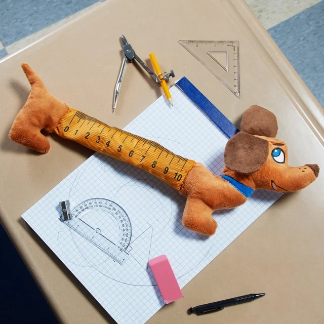 Overhead shot of dachshund plush toy doubling as a measuring tape, surrounded by geometric tools and placed on a desk.