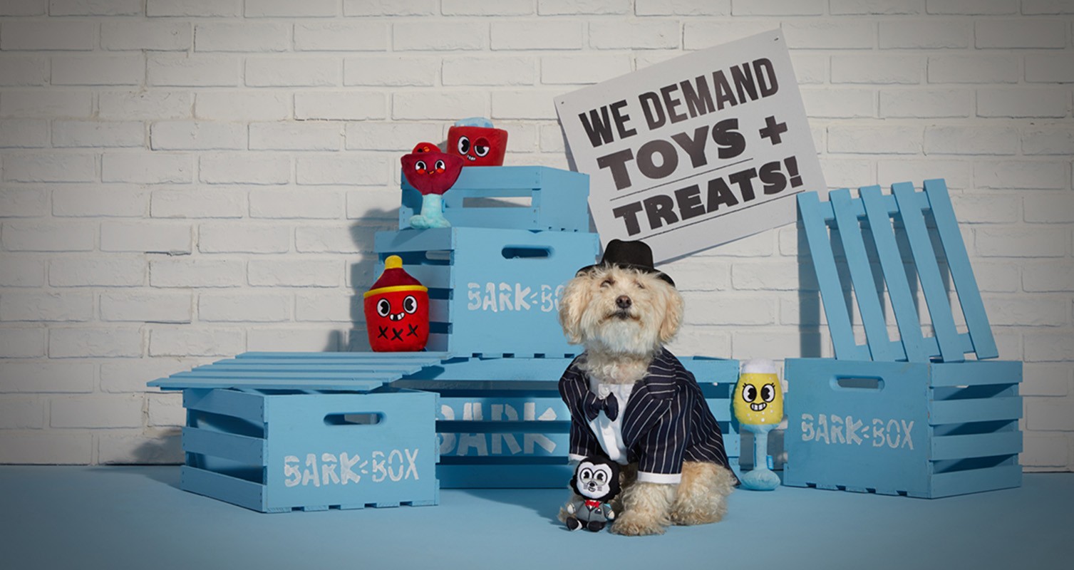 Dog in pinstripe suit and hat sits among blue crates with toys and a sign reading, 'We Demand Toys + Treats!' against a brick wall.