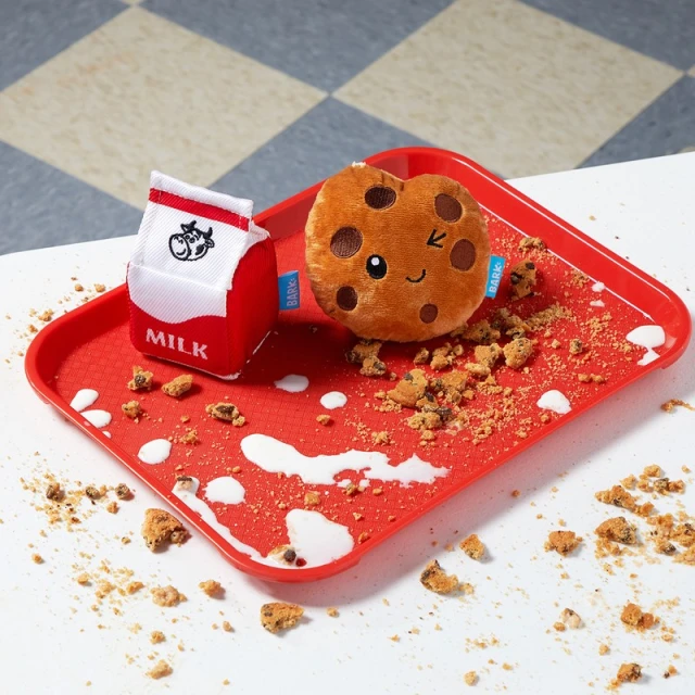 Stuffed milk carton and cookie toys are arranged on a red tray against a checkered floor with cookie crumbs and splashes of white liquid.