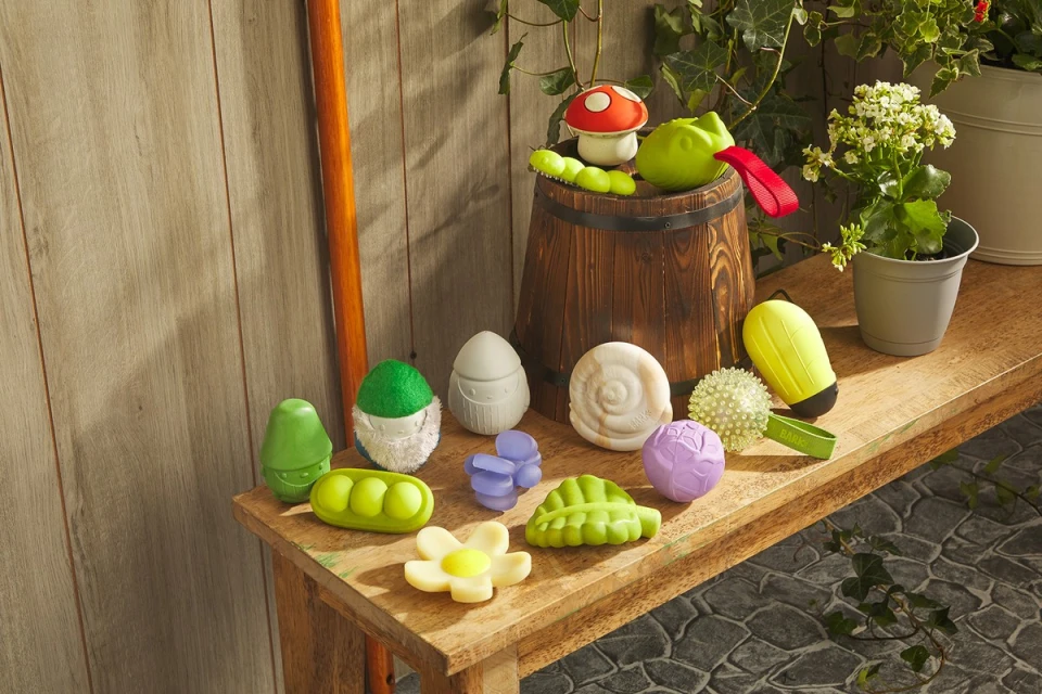 Still life of assorted colourful dog toys on wooden bench with barrel, flower pot, and foliage against panelled backdrop.