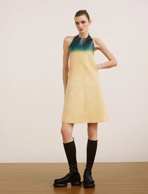 A slender female model showcases a halter dress in gradient yellow and teal, paired with knee-high socks and chunky black boots in a studio.