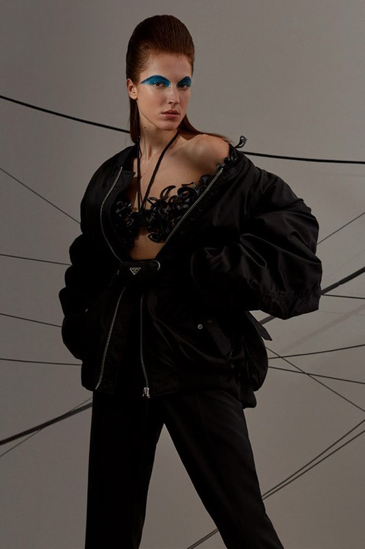 Model poses in black bra, pants, and unzipped bomber jacket against gray backdrop with thin black lines and dramatic blue eyeshadow.