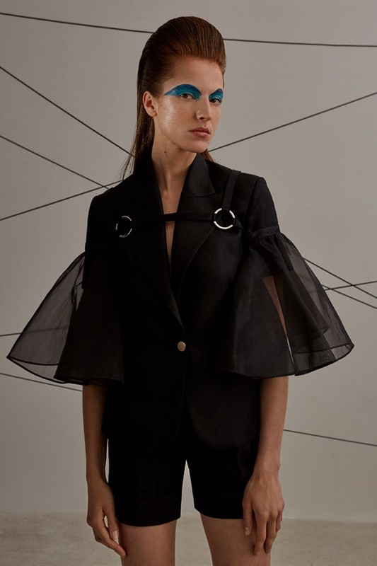 A woman with blue eye makeup poses in a black blazer with sheer sleeves and black shorts against a grey background with linear elements.