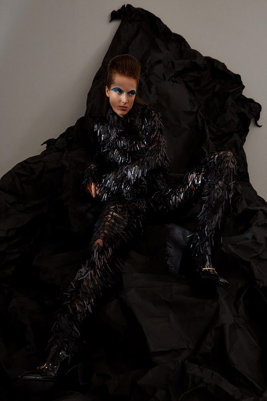 Woman in textured black outfit poses against draped black fabric. Shoulder-length dark hair, blue eyeshadow, studio lighting. Fashion-forward.