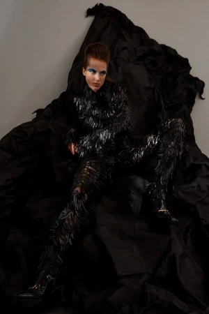 Woman in textured black outfit poses against draped black fabric. Shoulder-length dark hair, blue eyeshadow, studio lighting. Fashion-forward.