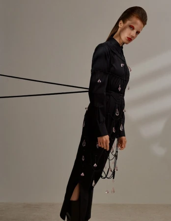 A model standing at a tilt in a black buttoned dress embellished with dangling pink crystal charms against a plain neutral background.