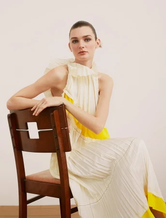 Model in a cream and yellow pleated dress seated backwards on a wooden chair against a plain light wall.