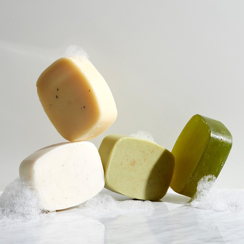 Four bars of natural soap in various colors are stacked on a reflective surface, surrounded by white foam against a neutral background.