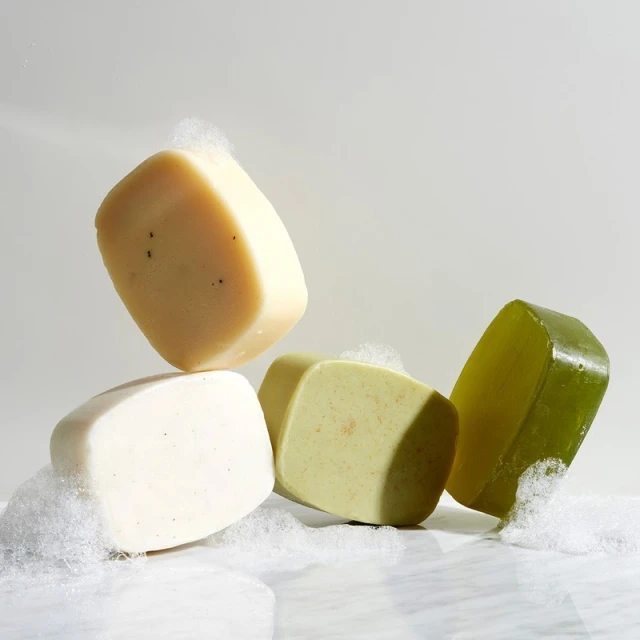 Four bars of natural soap in various colors are stacked on a reflective surface, surrounded by white foam against a neutral background.