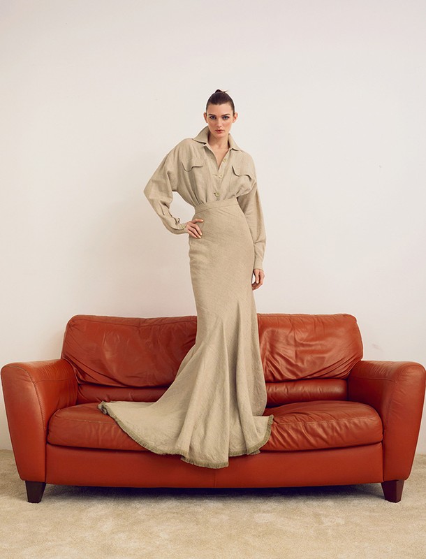 A full shot of a Caucasian fashion model posing on a burnt-orange couch in a light beige, long-sleeved shirt and tan, floor-length skirt.