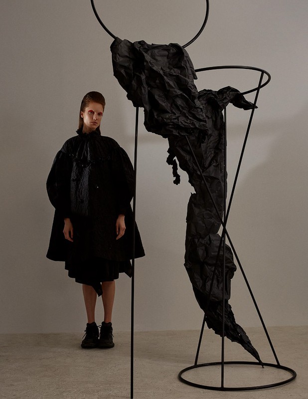 Avant-garde fashion shot of a poised adult female model next to a sculpture, against a neutral background in a photo studio.