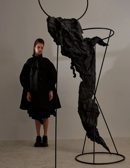 Avant-garde fashion shot of a poised adult female model next to a sculpture, against a neutral background in a photo studio.