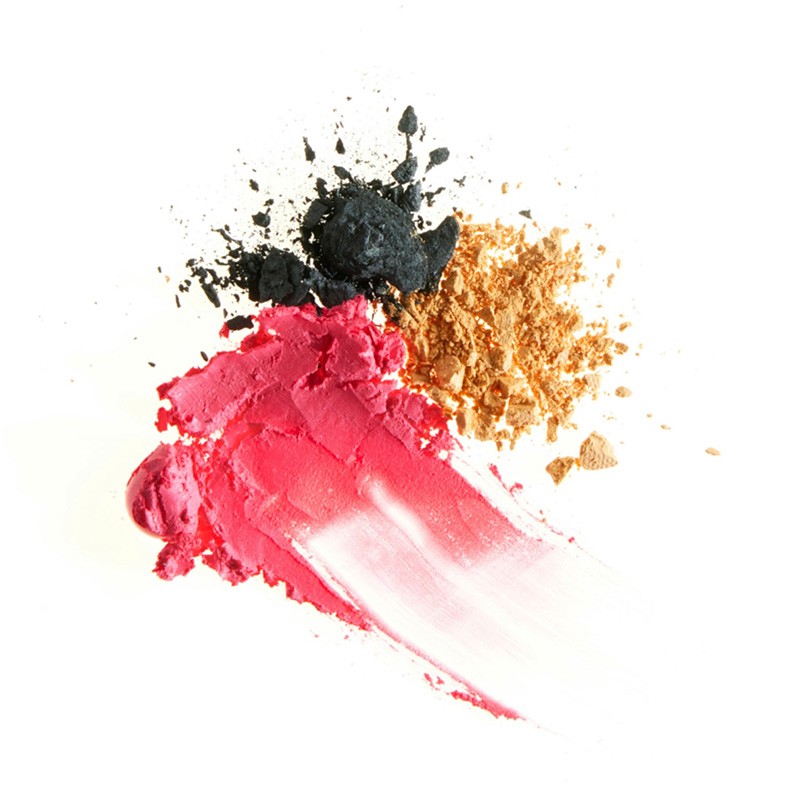 Cosmetics still life features smeared pink lipstick, black eyeshadow powder, and shimmering gold powder on a clean white surface.