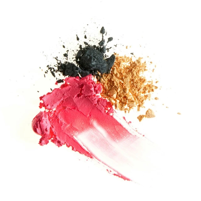 Cosmetics still life features smeared pink lipstick, black eyeshadow powder, and shimmering gold powder on a clean white surface.