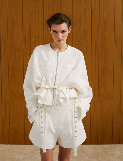 Fashionable woman in a studio setting poses in a textured white jacket and shorts set with a belt detail, standing against a brown wood paneled wall.