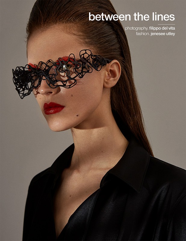 Close-up portrait of a woman wearing sculptural black lace-like eyewear and bright red lipstick against a plain grey background