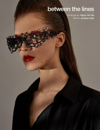 Close-up portrait of a woman wearing sculptural black lace-like eyewear and bright red lipstick against a plain grey background