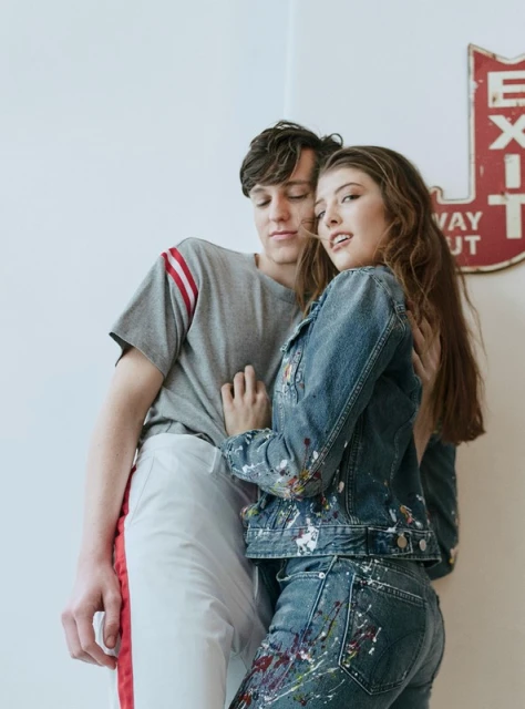 A young couple poses in front of a white wall with a vintage sign; the female wears a denim jacket with paint splatters, and the male wears a gray t-shirt.