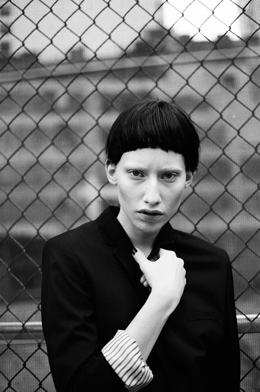 Model with short dark blunt bangs posing in a black blazer against a chain-link fence in Los Angeles