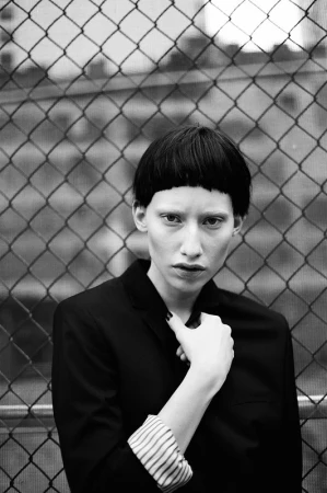 Model with short dark blunt bangs posing in a black blazer against a chain-link fence in Los Angeles