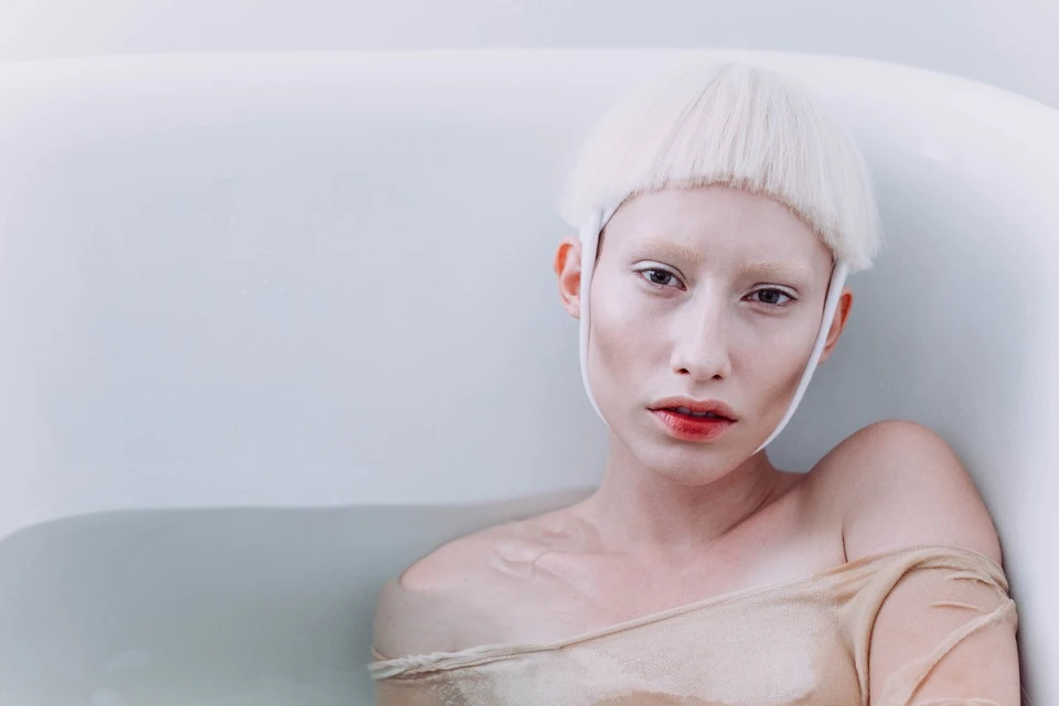 Portrait of an albino woman in a bathtub with off-the-shoulder top, showcasing pale skin, light hair, and red lipstick in a minimalist setting.