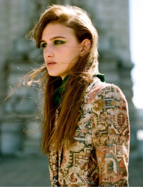 Woman with auburn hair and green eyeshadow poses in a patterned jacket against an architectural background on a breezy day.