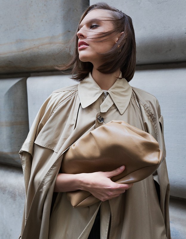 Fashion shot of an adult female in a trench coat holding a leather clutch against a concrete wall, medium shot, wind-blown hair.