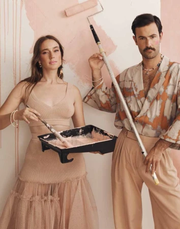 A couple in stylish attire is painting a white wall with blush pink paint. The woman holds a paint tray, and the man holds a paint roller.