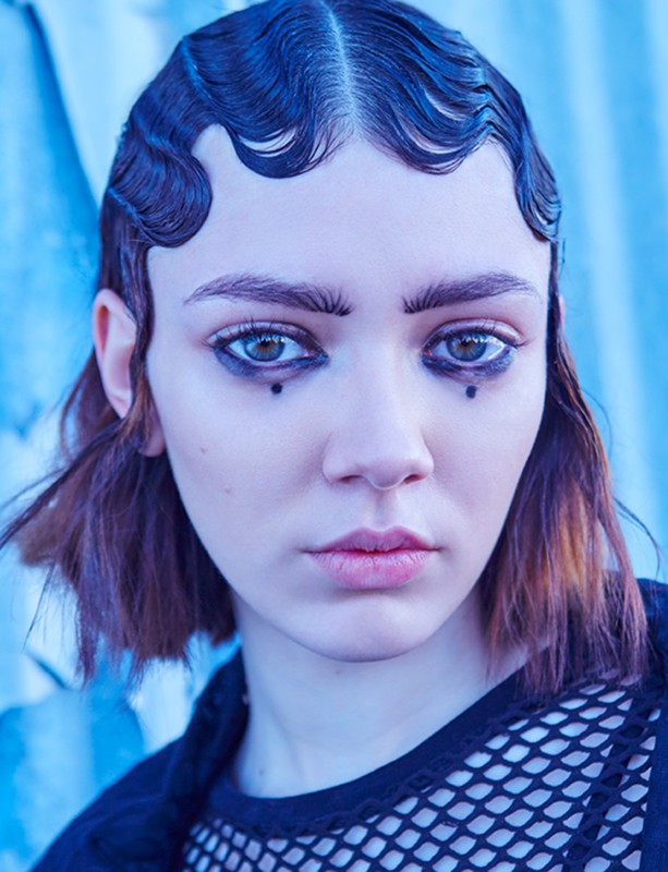 Portrait of a young woman with artistic makeup and distinctive hairstyle against a blurred blue background in a beauty or fashion context.