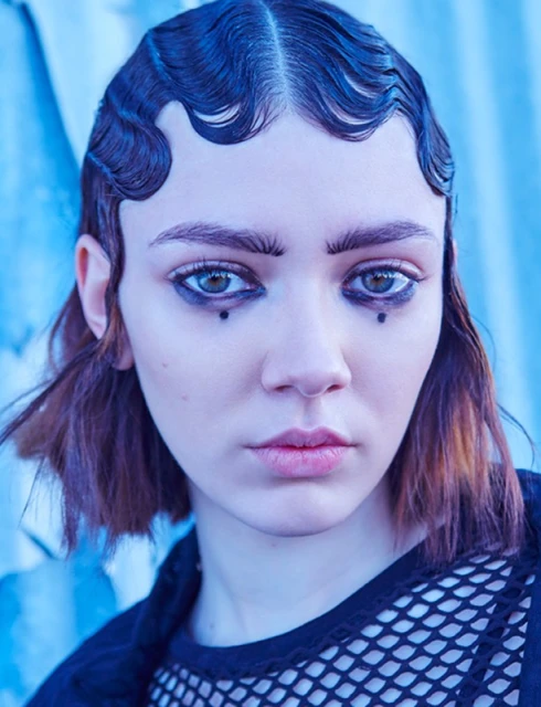 Portrait of a young woman with artistic makeup and distinctive hairstyle against a blurred blue background in a beauty or fashion context.