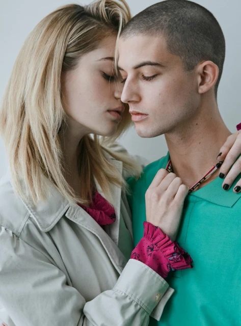 Fashion-style close-up of a blonde woman and a man with short hair, embracing in front of a white background, wearing colorful tops and a beige coat.