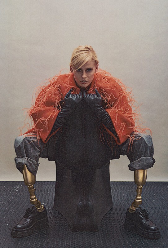 Fashion editorial shot of a woman with prosthetic legs sitting on a black sculpture. She is wearing an orange feathered coat and jeans.